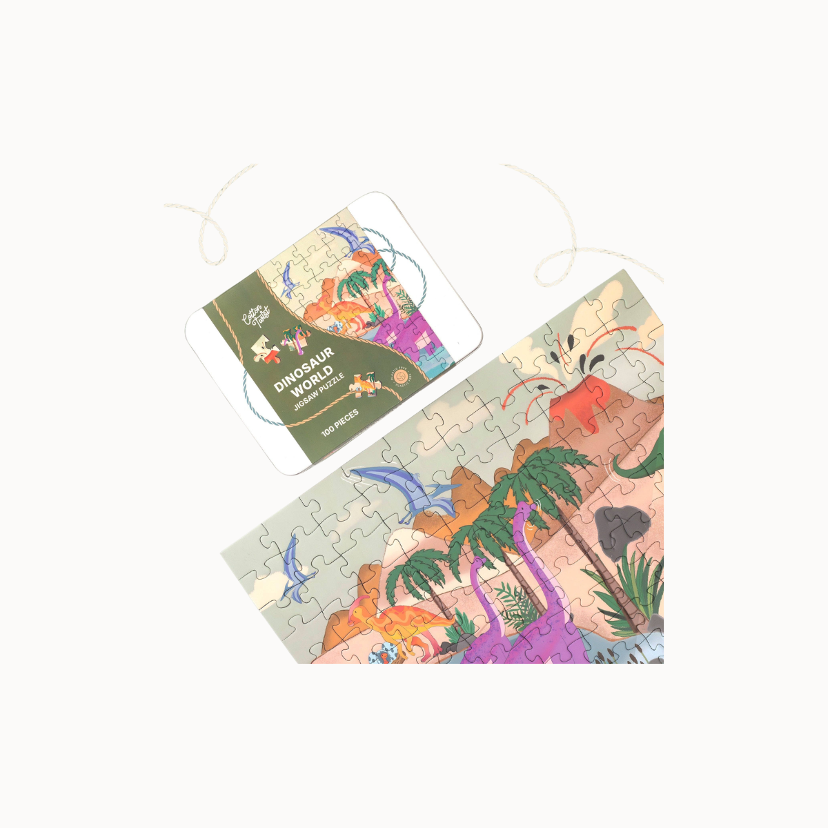A children's jigsaw puzzle and its matching cover card featuring a colorful dinosaur landscape with palm trees, mountains, and a blue bird, shown on a plain light background.