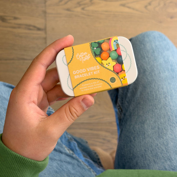 Hand holding a small tin labeled 'Good Vibes Bracelet Kit' with colorful designs on a wooden floor.