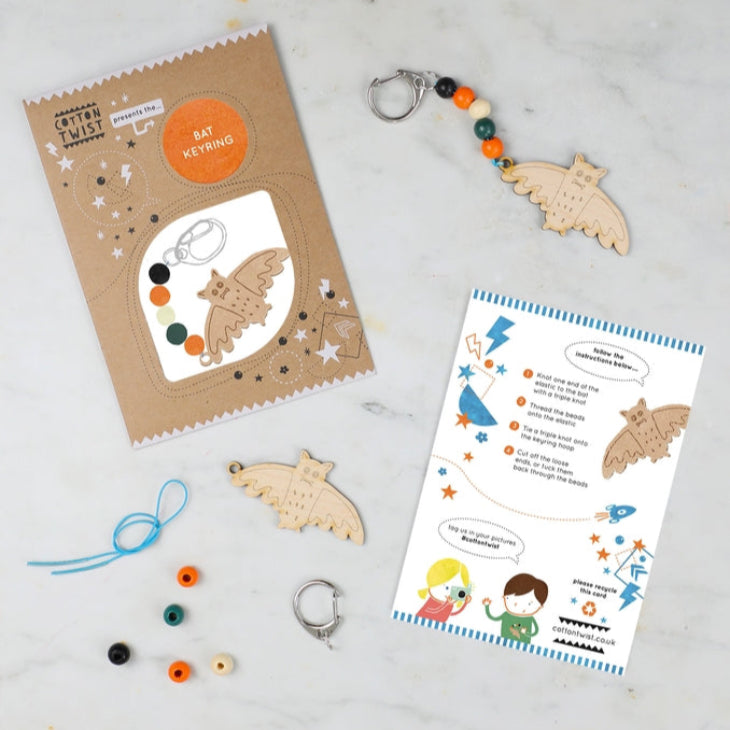 A craft kit for making a Halloween-themed keyring, including a maple wood bat-shaped fob, colorful beads, an elastic with metal ends, and a printed instruction card, all displayed on a marble surface.