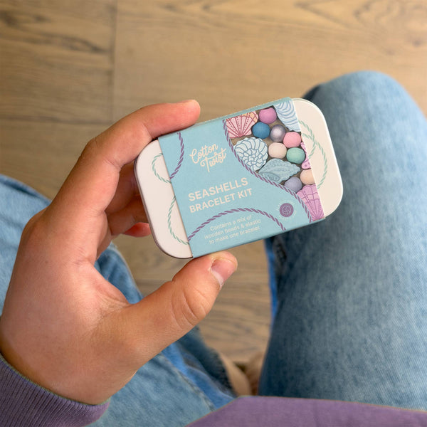 Hand holding a small tin labeled 'Seashells Bracelet Kit' on a wooden floor.