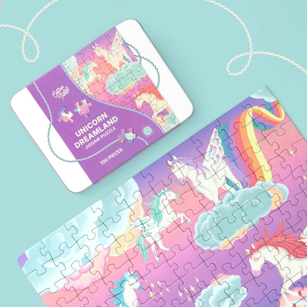 A colorful unicorn-themed jigsaw puzzle and its matching puzzle box are displayed on a light aqua background with decorative rope loops. The box reads 'Unicorn Dreamland Jigsaw Puzzles' and shows pastel illustrations of unicorns, castles, clouds, and a rainbow.