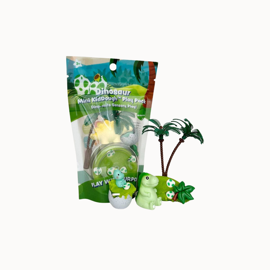 Mini McDough Dinosaur Play Pad set in its packaging, featuring a green dinosaur egg, a light green dinosaur figure, two small palm trees, and a soccer ball on a plain white background.