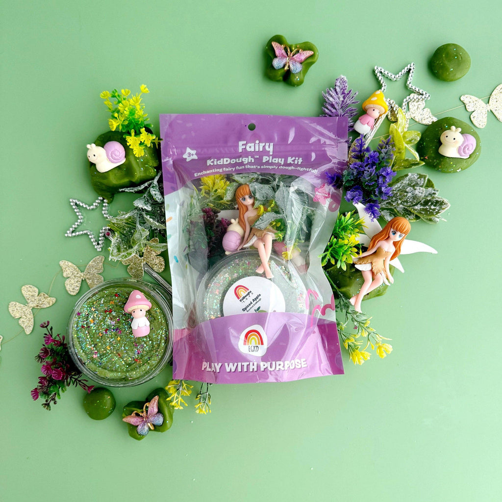 A packaged fairy-themed play kit sits on a mint green background surrounded by decorative craft props, including glitter, butterflies, stars, flowers, and small fairy figurines. The central product is a clear plastic bag with a purple header reading 'Fairy KidDough Play Kit.