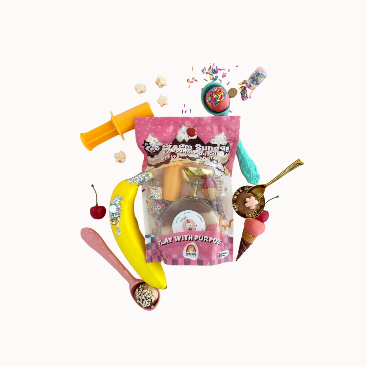 A colorful toy set arranged around a pink bag labeled 'Play with Purpose' on a plain white background. The set includes a yellow toy banana, a pink spoonful of beads, a small cherry, a brown ice cream cone toy, a yellow syringe-like tool, and other small accessories.