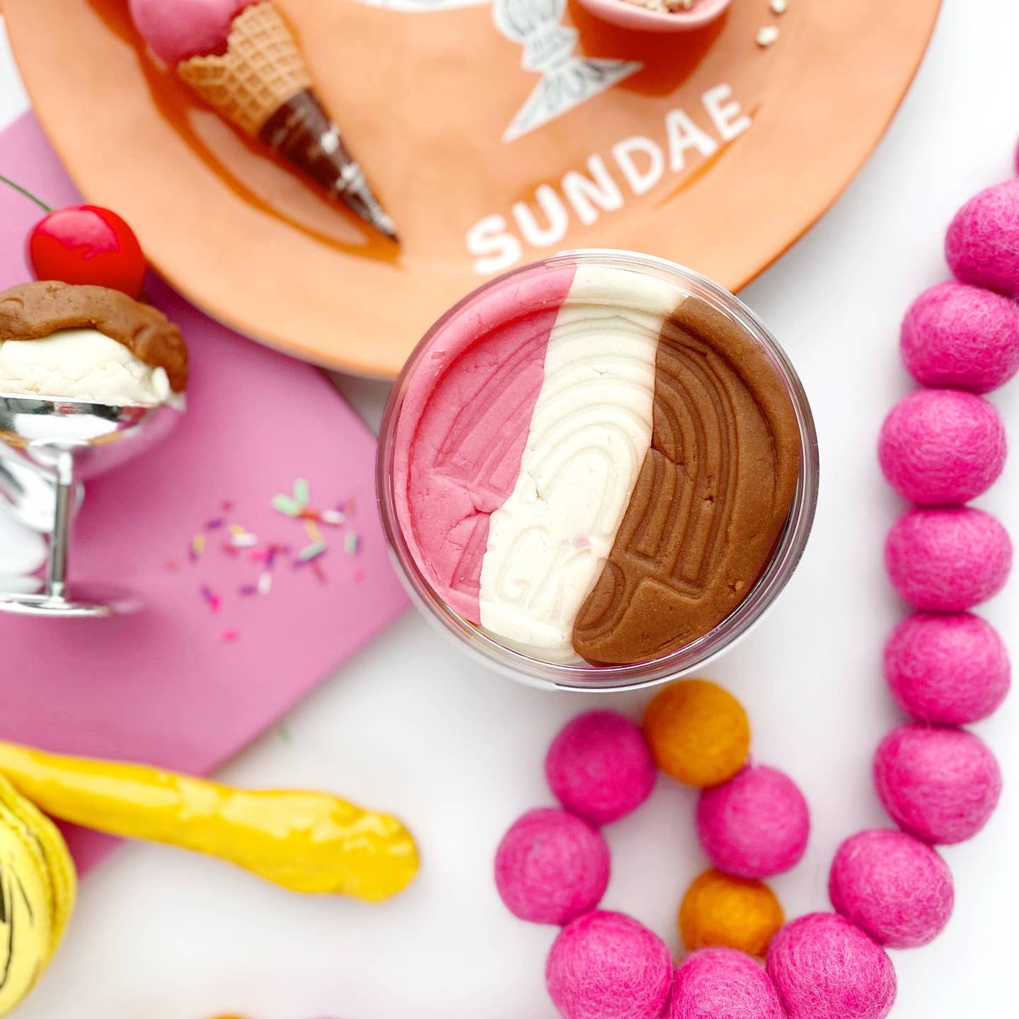 A top-down styled shot of a dessert sundae in a clear glass cup, with pink, white, and brown swirls. Surrounding props include a pink plate with the word SUNDAE, pink felt beads, a yellow toy, and ice cream dishes on a white surface.