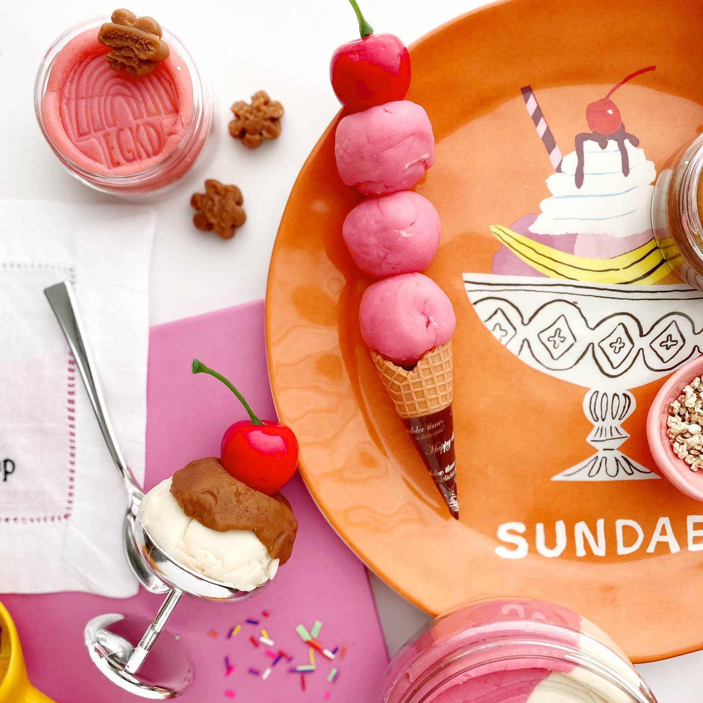 Ice cream cone with pink ice cream on an orange plate labeled 'Sundae' with cherry and cookie decorations.