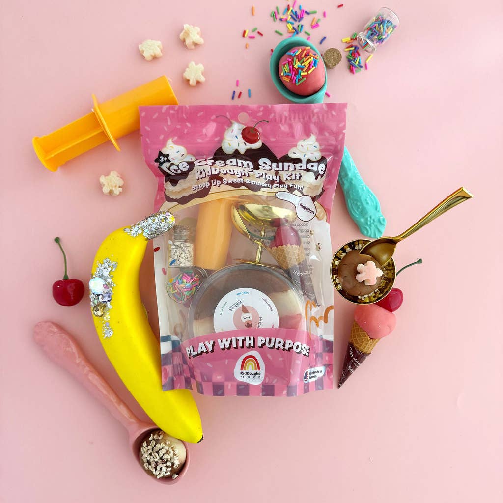 An ice cream sundae-themed play with purpose toy kit arranged in a flat lay on a pink background. The central product is a pink bag labeled 'Ice Cream Sundae Play With Purpose' containing a pretend sundae dish, a bowl, and accessories.