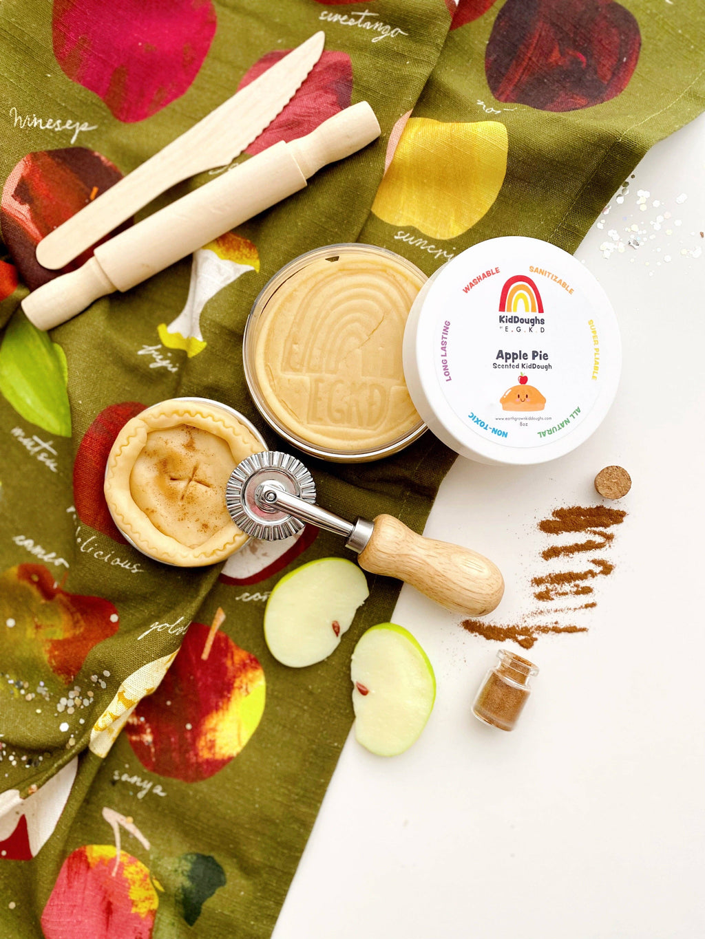 Apple pie playdough kit with a rolling pin, apples, and a decorative cloth on a white surface.