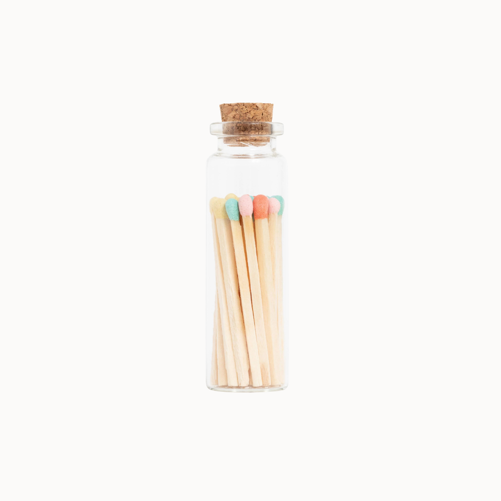 A small clear glass bottle with a cork stopper containing wooden matches with pastel-colored tips, centered on a plain white background.