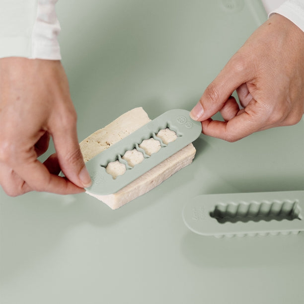 Two gray silicone food cutters designed for baby-led weaning, one for creating finger-length strips and the other for bite-sized cubes.