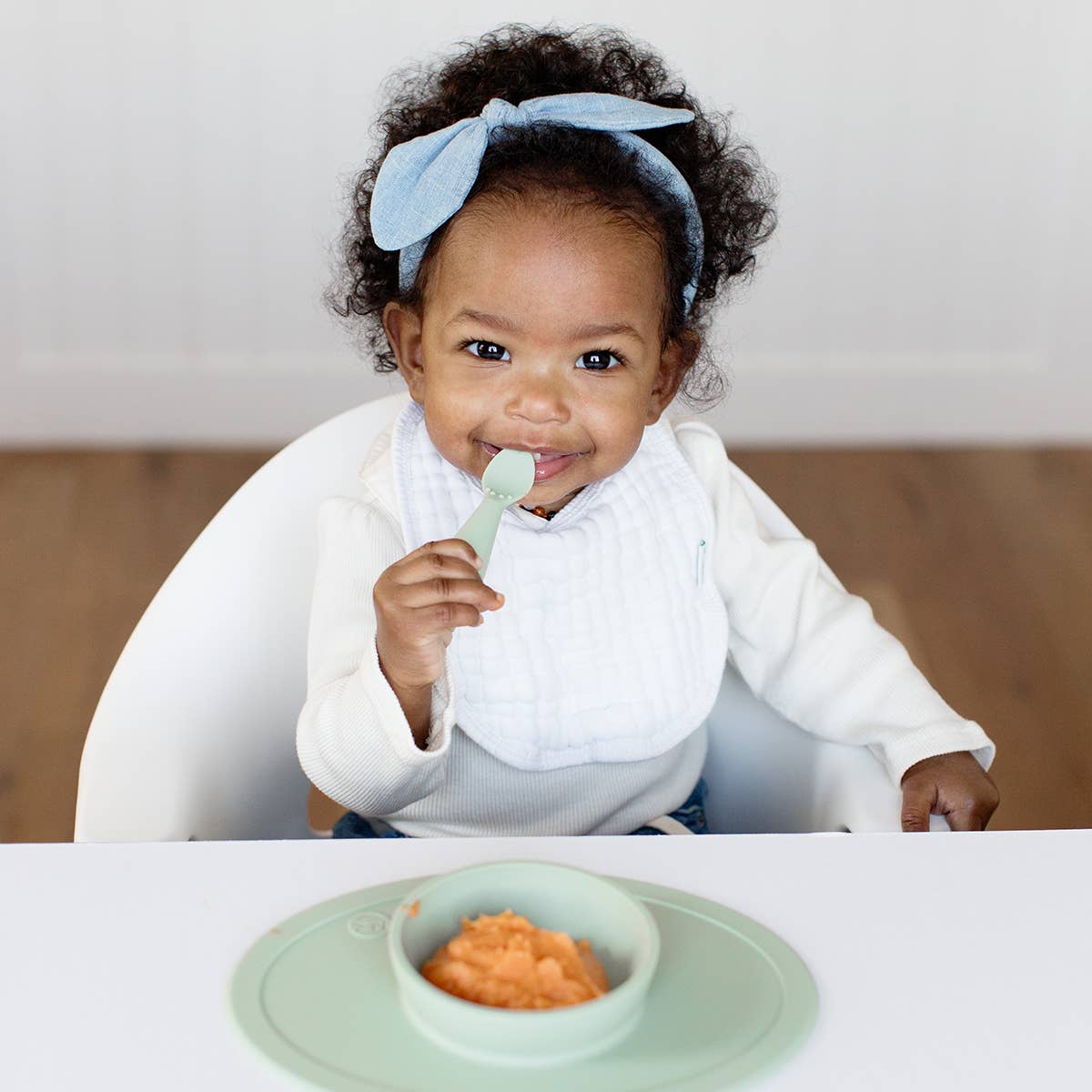 Baby sitting in a high chair with a bowl of food and a spoon, wearing a white bib and blue headband.