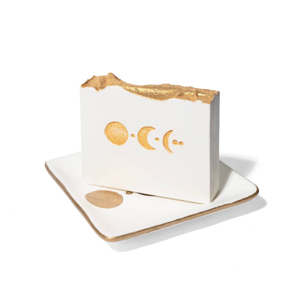 A white rectangular soap bar with gold foil moon phases on the front, standing on a matching white square dish with a gold rim against a white background.