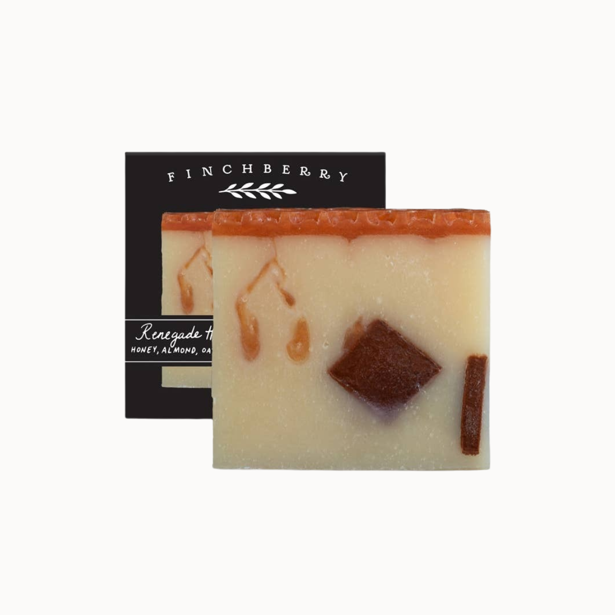 A square handmade soap bar with a cream-colored base, a terracotta orange top layer, and botanical inclusions, shown in front of its black packaging box labeled FINCHBERRY.