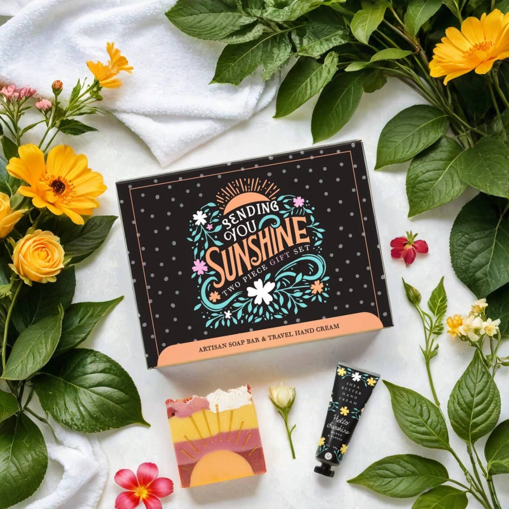 Gift set with 'Sending You Sunshine' packaging, soap bar, and hand cream surrounded by flowers and leaves on a white background.