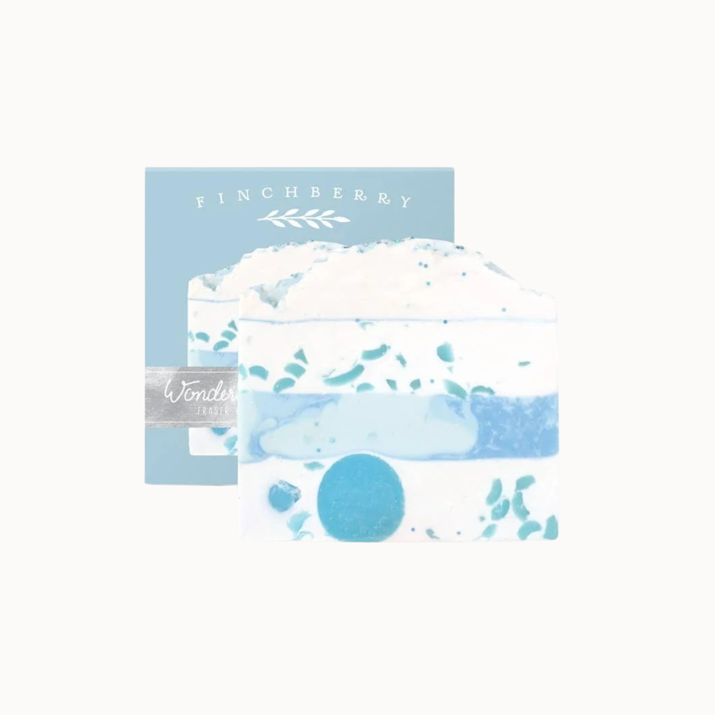 A layered artisanal soap bar in white and light blue with teal leaf accents, shown in front of its light blue packaging box labeled 'FINCHBERGY' and 'Wonders'.