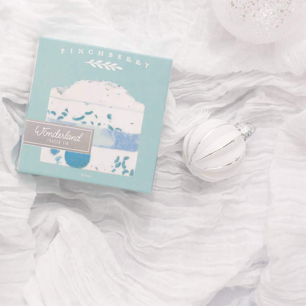 A boxed bath bomb set labeled Finchberry Wonderland Magic Pie sits on white draped fabric next to a white ornament.