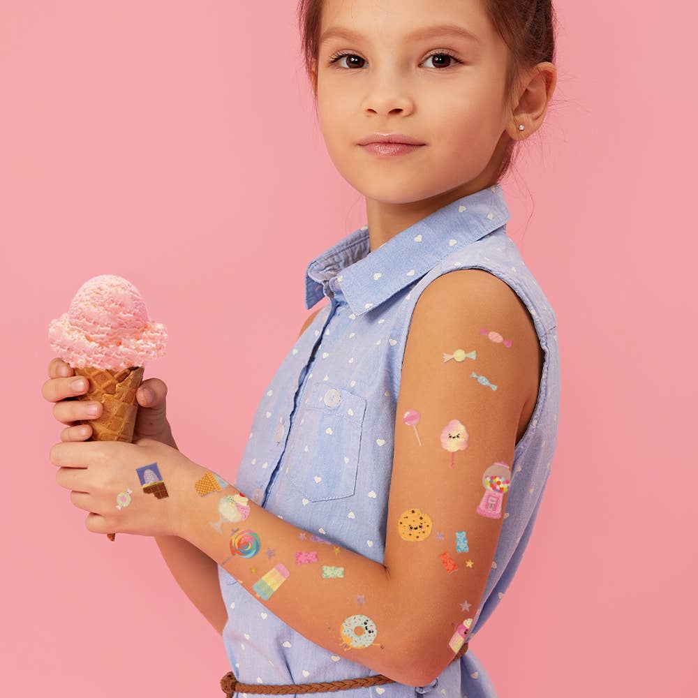 Young girl holding a pink ice cream cone with temporary tattoos on her arm against a pink background