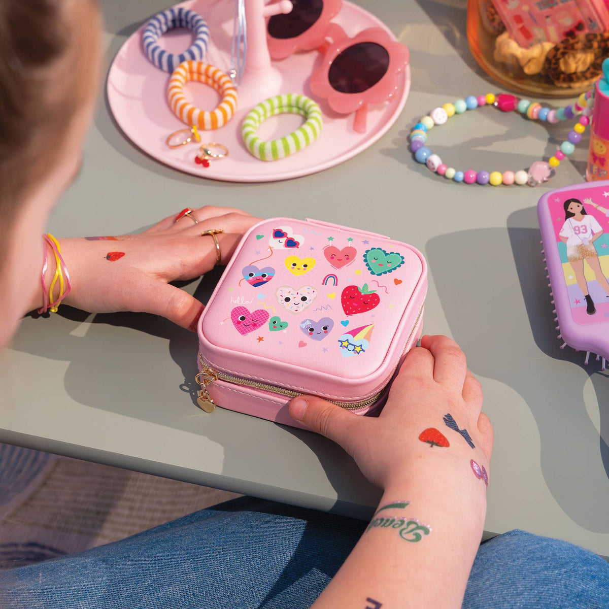 A child's hands hold a small pink zippered case decorated with colorful cartoon stickers, set in a lifestyle context with other kids' accessories on a table.