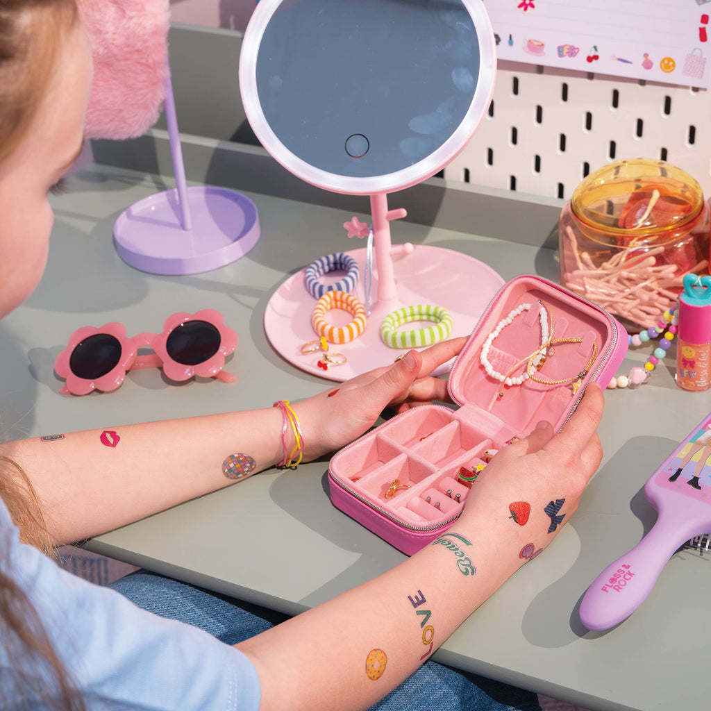 A child's hands with temporary tattoos hold an open pink jewelry organizer case containing various necklaces and bracelets. The scene is set on a gray desk with a pink vanity mirror, pink sunglasses, and other colorful accessories.