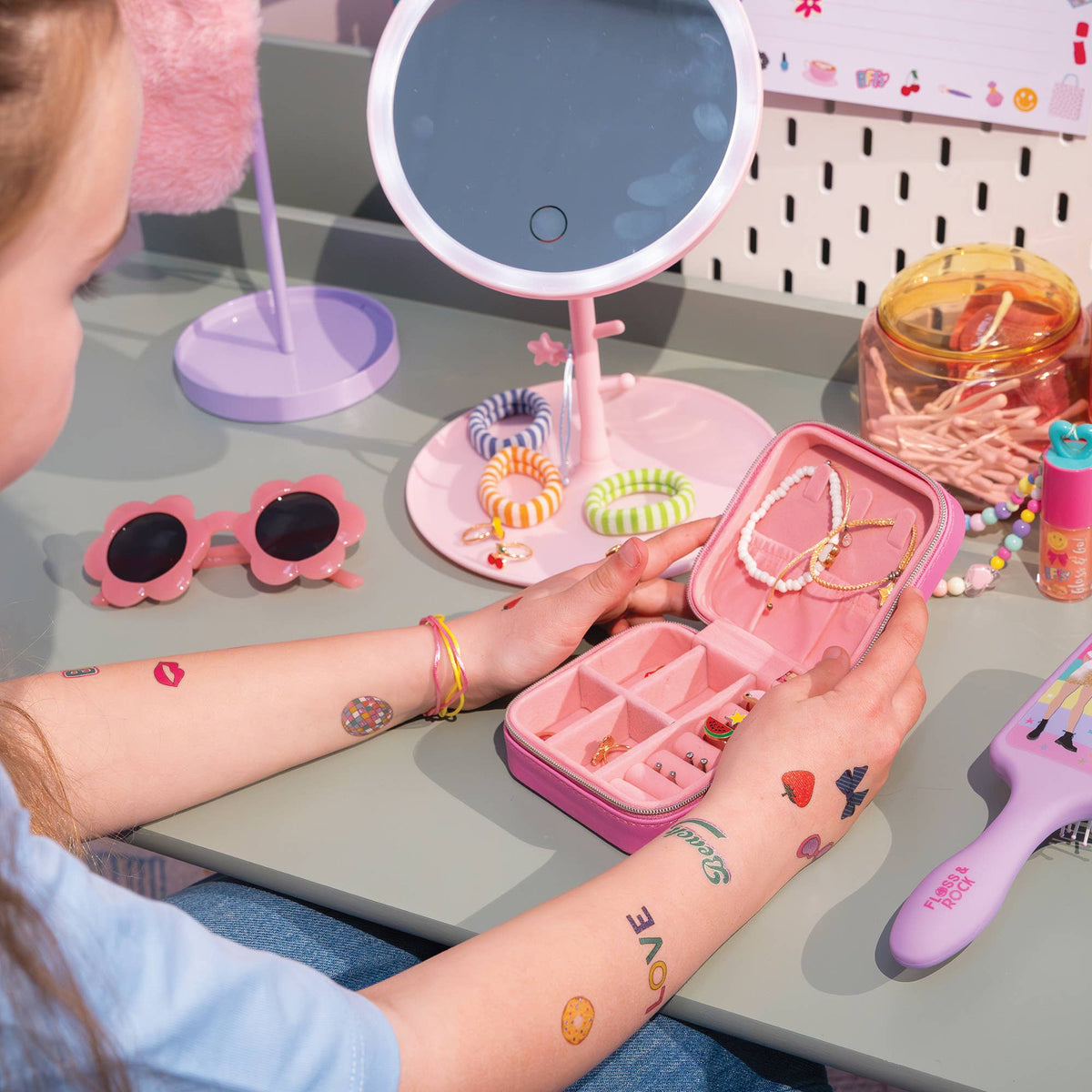 A child's hands with temporary tattoos hold an open pink jewelry organizer case containing various necklaces and bracelets. The scene is set on a gray desk with a pink vanity mirror, pink sunglasses, and other colorful accessories.
