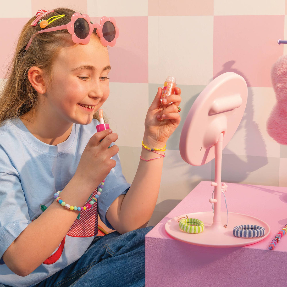A young girl with pink sunglasses on her head applies lip balm from a pink tube while looking into a pink vanity mirror. She is wearing a light blue t-shirt and jeans, with colorful hair clips and bracelets, in a pastel pink and white checkered room.