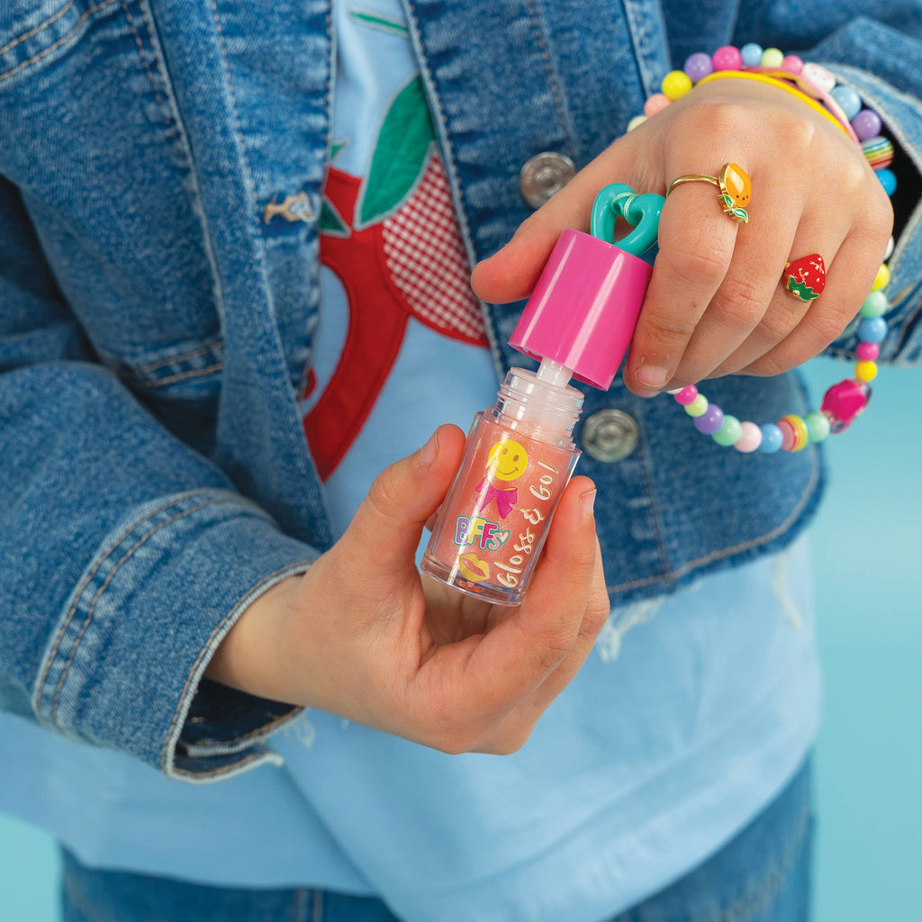 A close-up of a person's hands holding a small clear bottle of pink lip gloss with a bright pink cap and a teal heart-shaped applicator. The person is wearing a denim jacket, a light blue graphic T-shirt, and colorful beaded bracelets and rings against a solid light blue background.