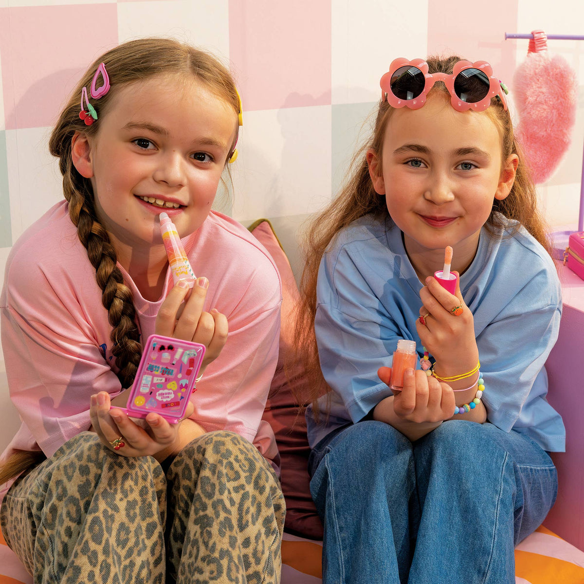 Two young girls sitting side-by-side holding pink toy cosmetics, with a pink and white checkered wall in the background.