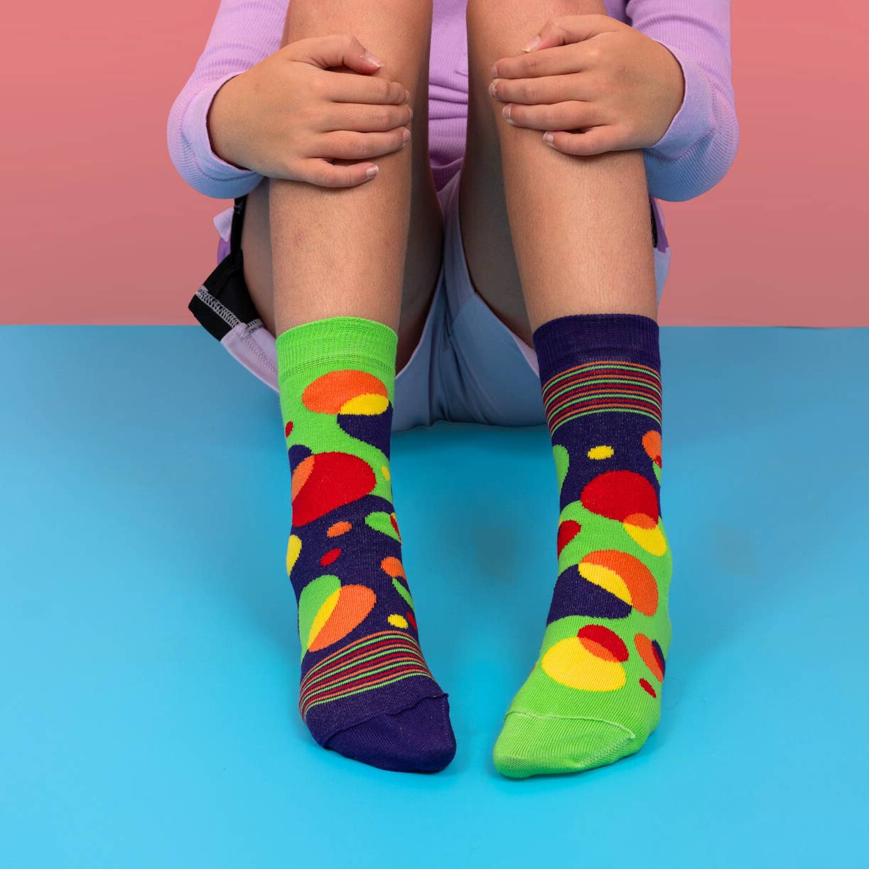Colorful socks with abstract patterns worn by a person sitting on a blue and pink background