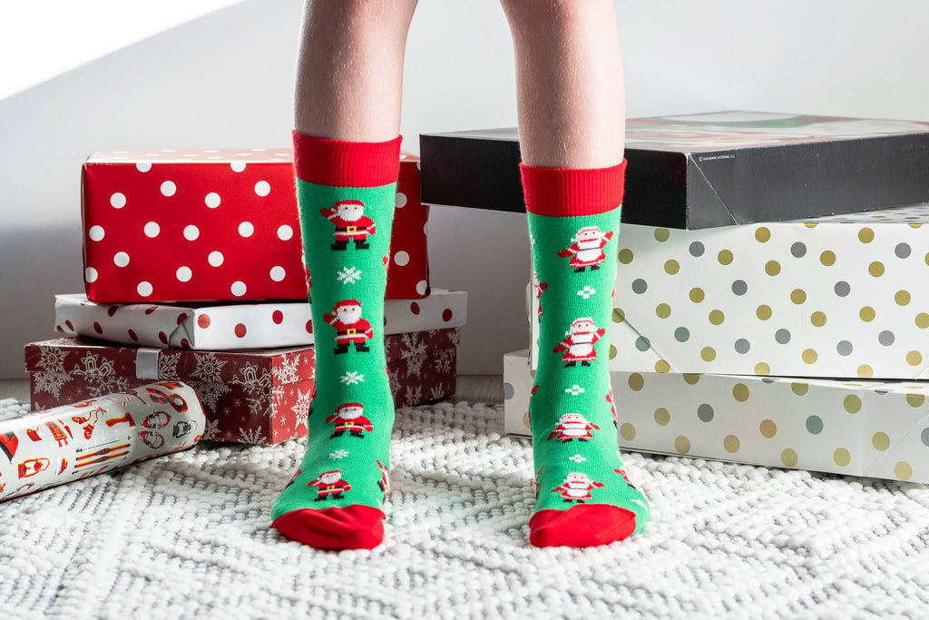 A person wearing bright green Christmas crew socks with red cuffs, heels, and toes, printed with Santa Claus and snowflake motifs. The person is standing on a white textured rug surrounded by wrapped gift boxes and presents.