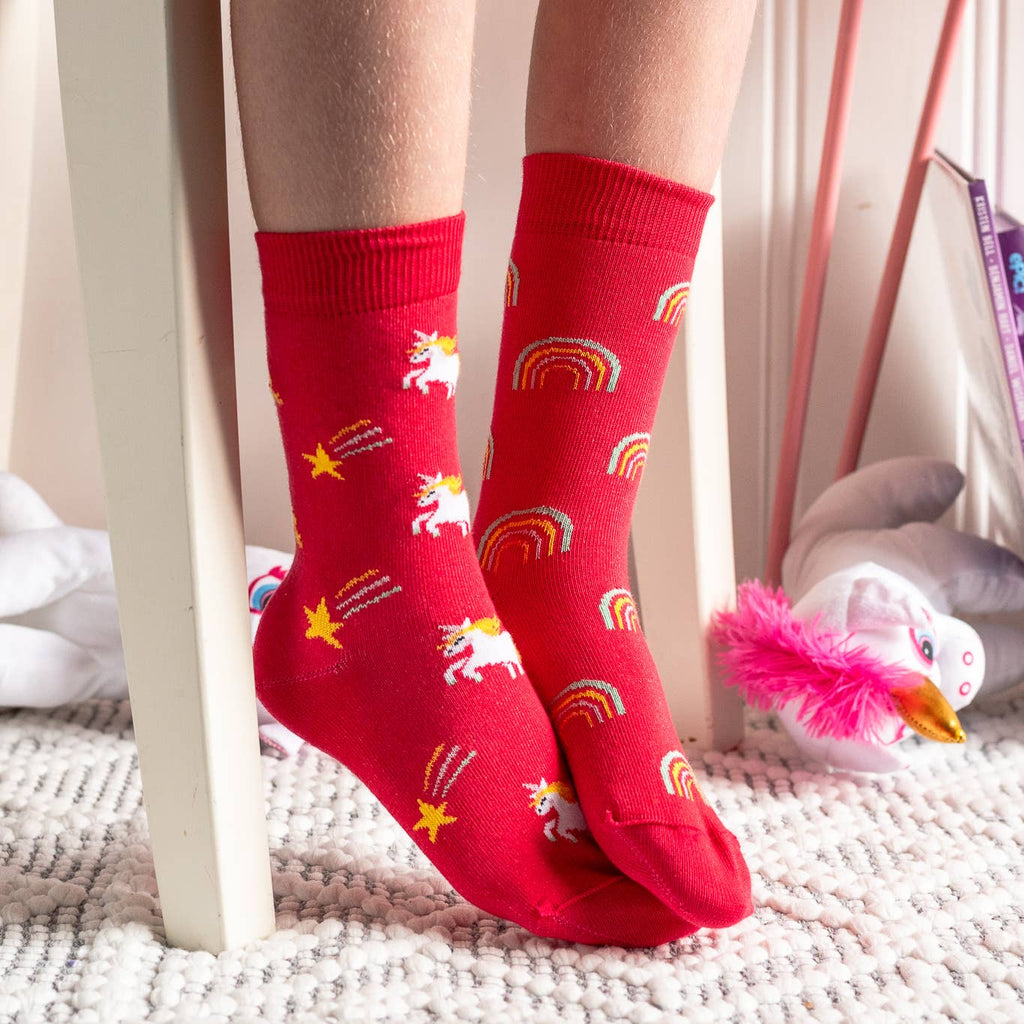 Red socks with unicorn and rainbow patterns worn by a person sitting on a bed.