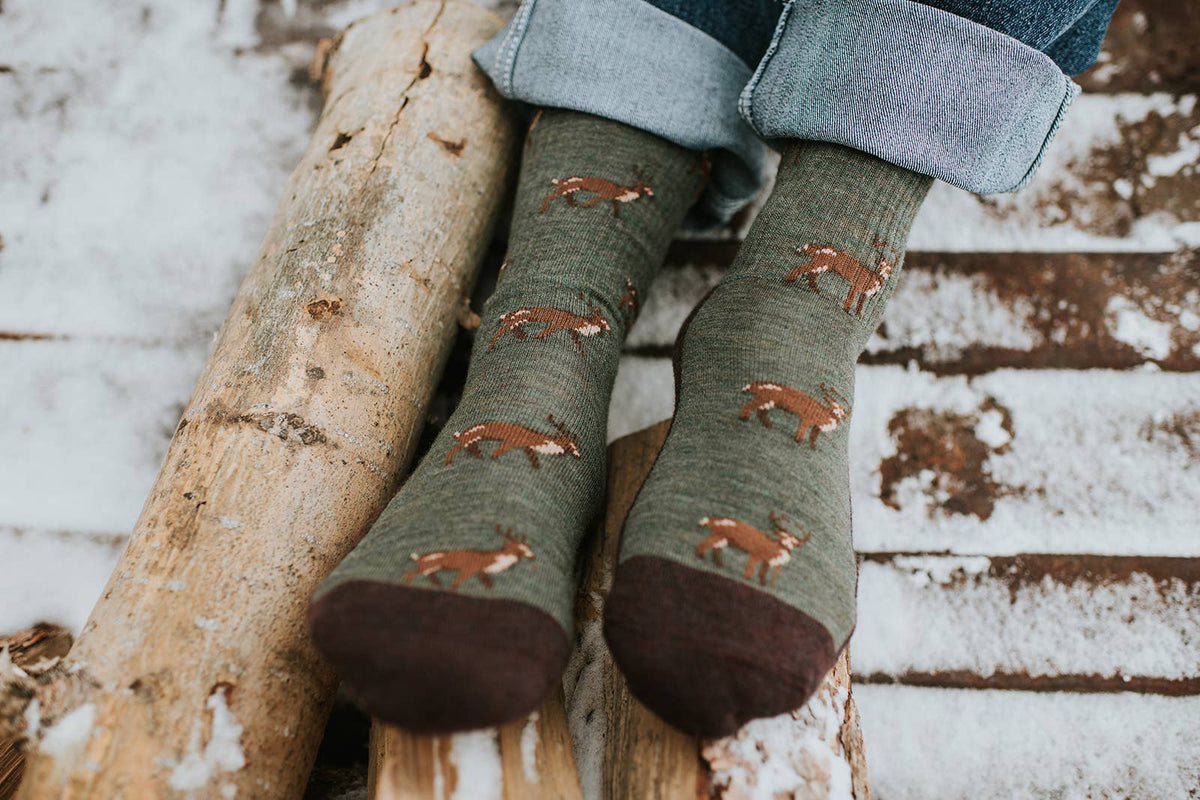 Green socks with deer pattern worn by a person sitting on a log in a snowy setting