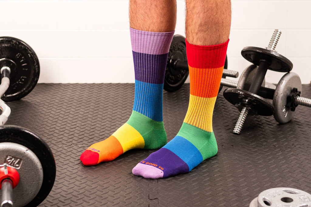 A person wearing rainbow-striped crew socks standing on a textured gym floor with dumbbells visible in the background.