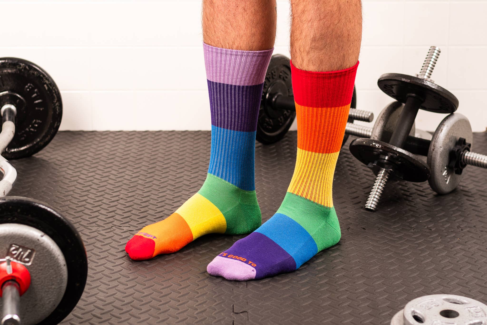 A person wearing rainbow-striped crew socks standing on a textured gym floor with dumbbells visible in the background.