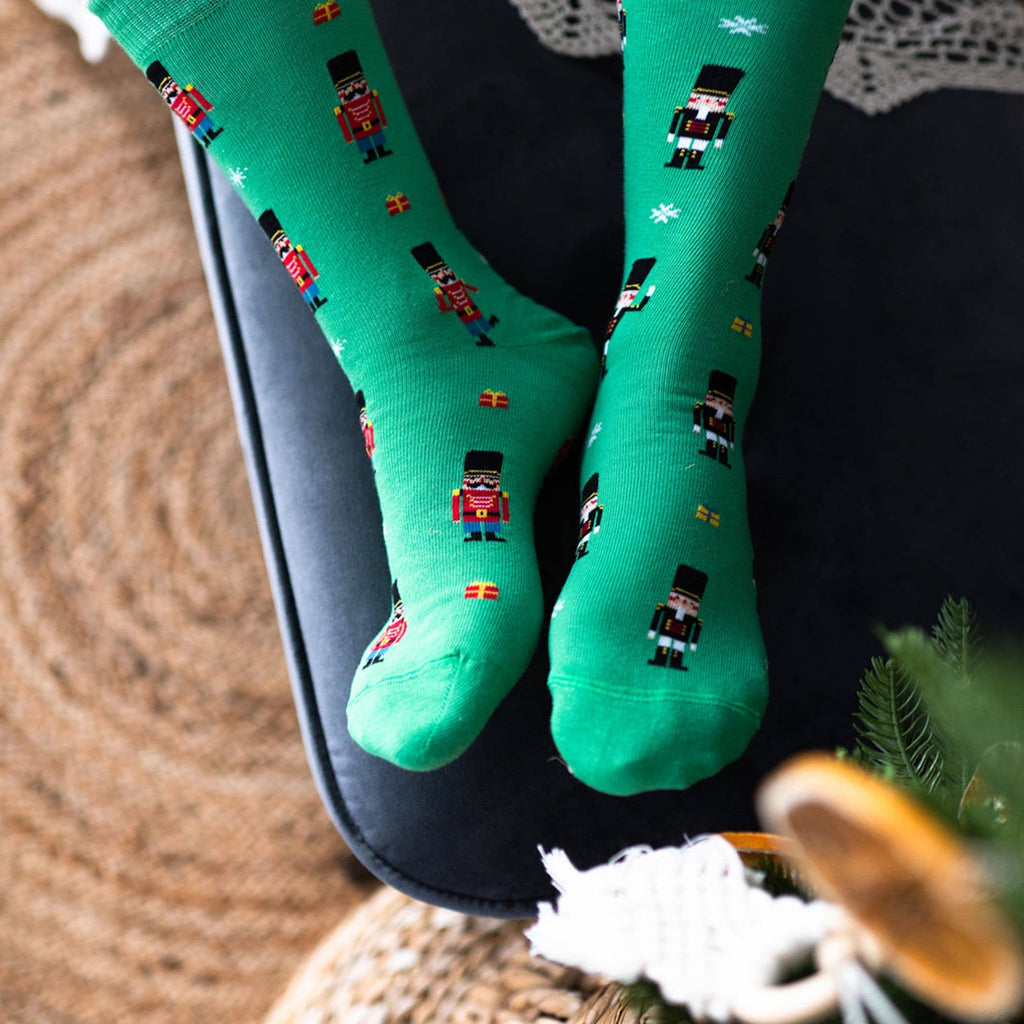 A pair of bright green crew socks with a repeating pattern of pixel-art nutcracker soldiers, worn on feet resting on a black chair.