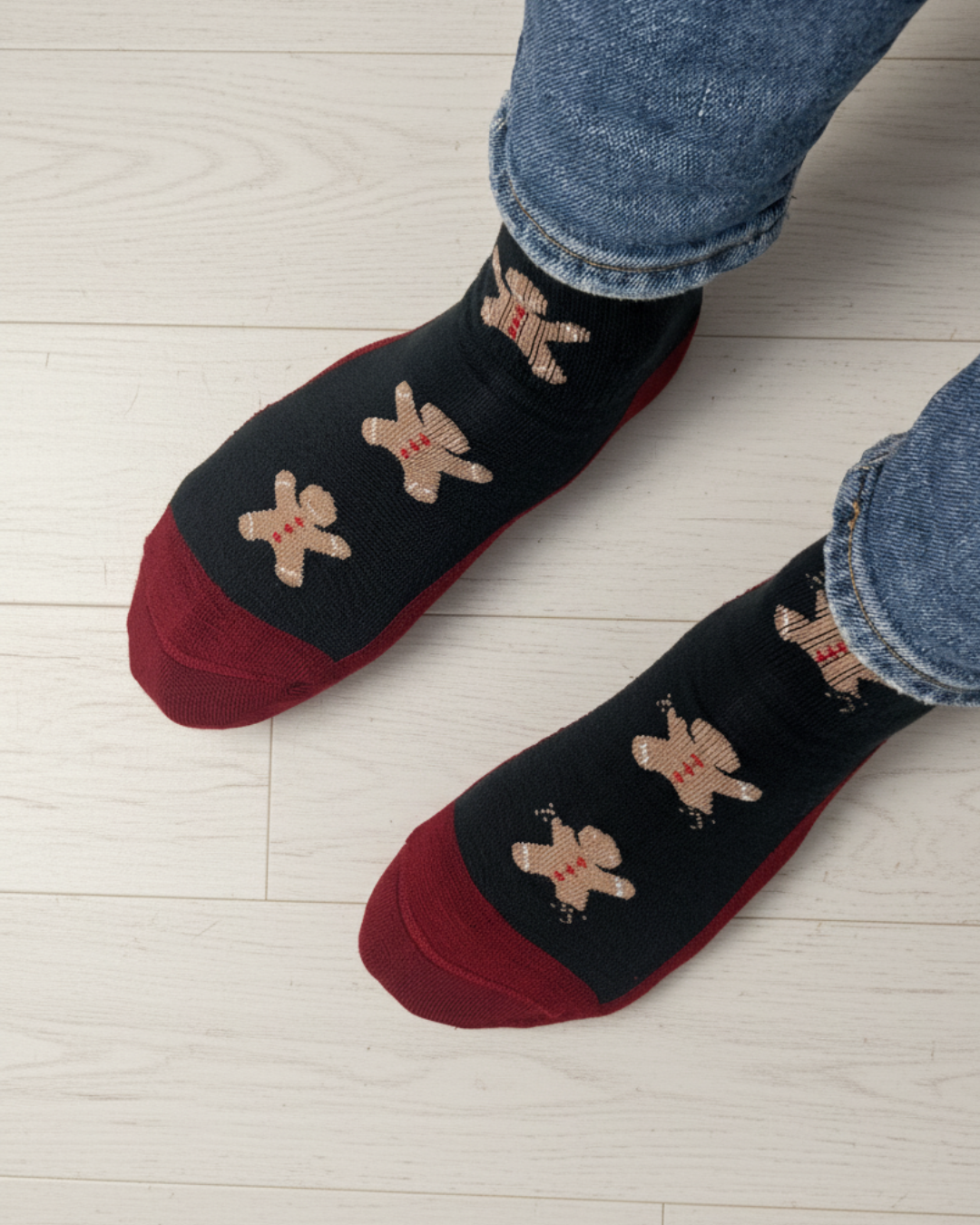 A pair of black crew socks with red toes and heels, decorated with beige gingerbread men motifs, worn with blue jeans on a light wood floor.