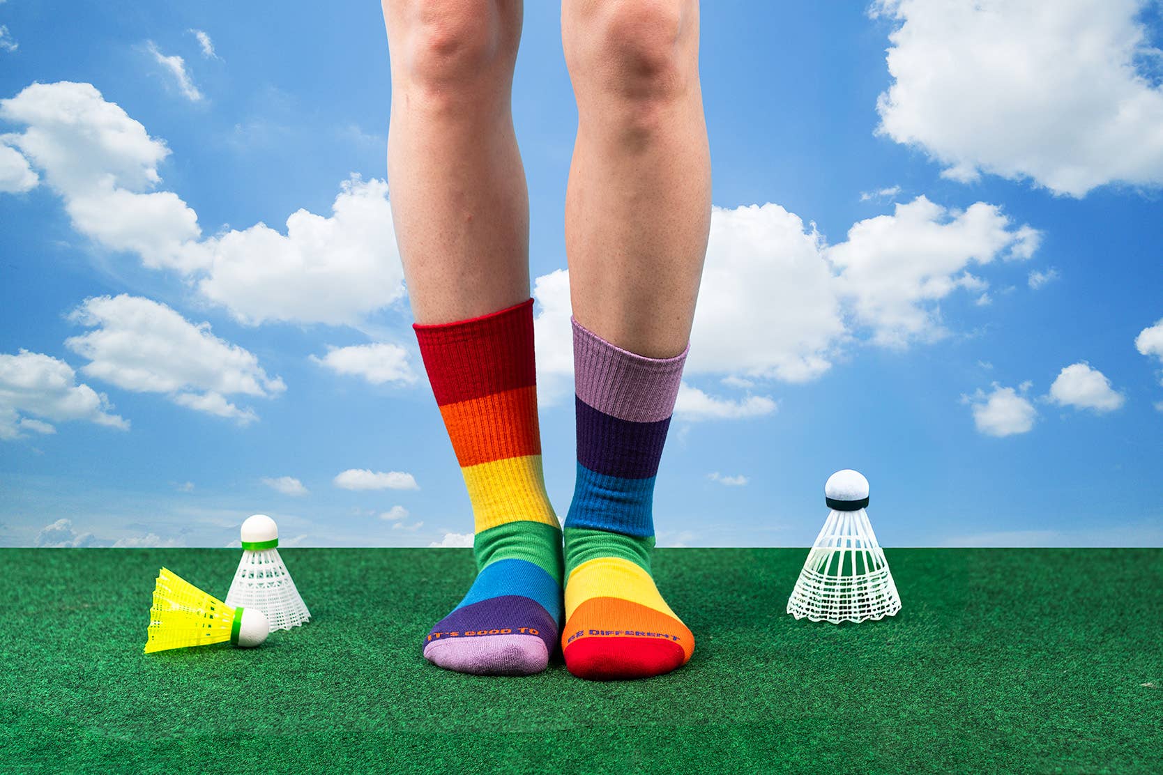 Colorful socks on feet standing on a green surface with badminton equipment and a blue sky.