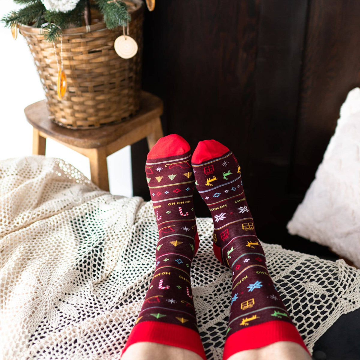 A pair of feet wearing dark red Christmas-themed crew socks with colorful holiday motifs and red cuffs, styled in a cozy indoor setting with a lace blanket, wooden furniture, and a basket in the background.