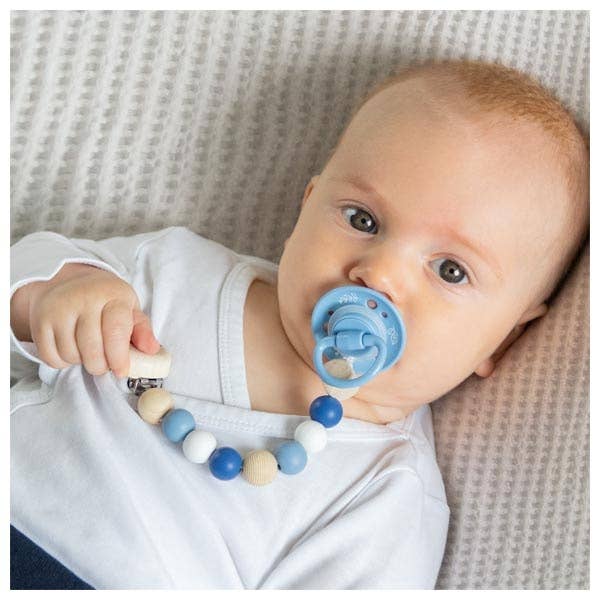 Baby holding a teething ring with blue pacifier on a textured surface