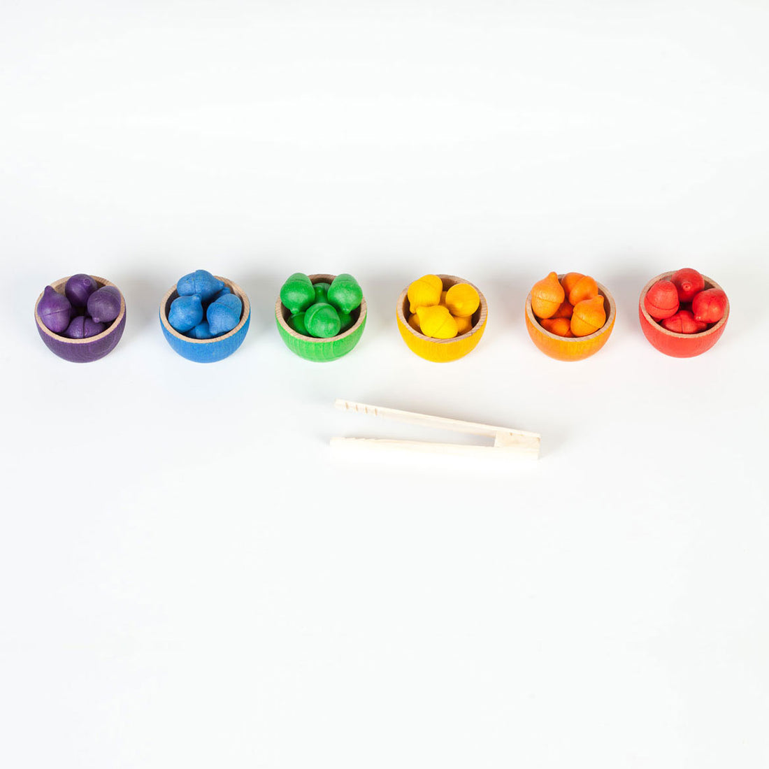 A set of six small wooden bowls, each painted in a different rainbow color and filled with matching round fruit-like pieces, arranged in a row on a white background with a pair of wooden tongs below.