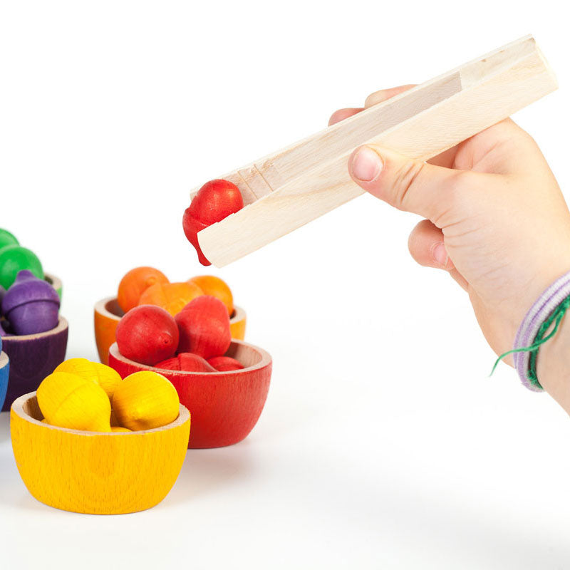 A hand holds a wooden toy scoop over small wooden bowls filled with colorful round fruit-like pieces on a white background.