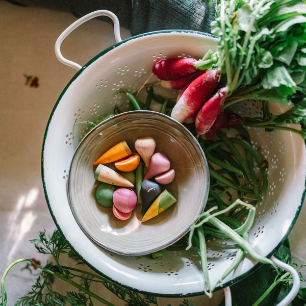 Colander with wooden vegetables and a bowl of vegetables on a wooden surface