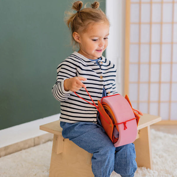 Child sitting on a small stool holding a pink backpack in a classroom setting.