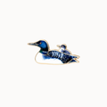 A loon bird enamel pin featuring a large blue and black loon with a smaller matching loon chick riding on its back, outlined in gold metal against a plain off-white background.