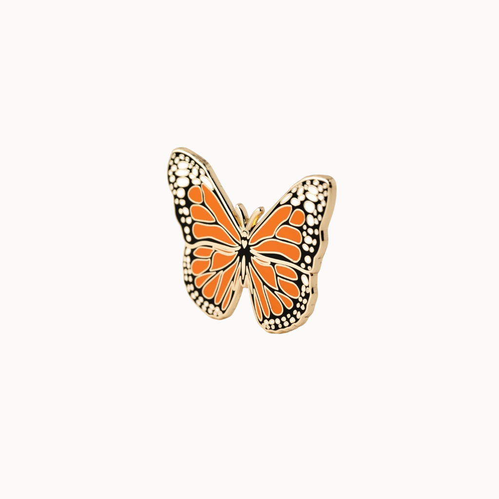 A butterfly-shaped enamel pin with orange wings, black and white spotted borders, and a gold-tone metal outline, centered on a plain off-white background.