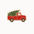 A festive enamel pin depicting a red vintage pickup truck carrying a green Christmas tree with gold and red ornaments, set against a plain white background.