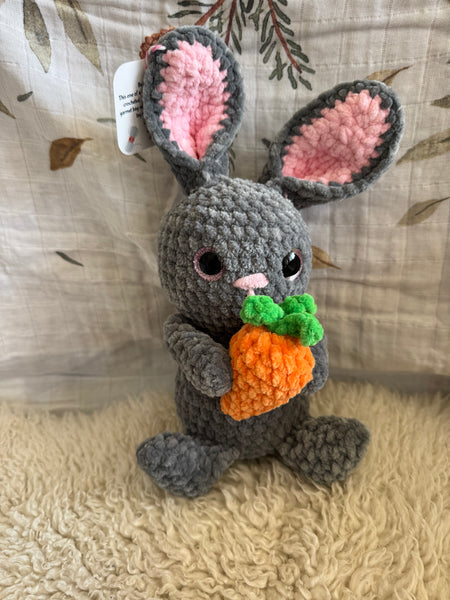 Gray plush bunny toy resembling a rabbit with pink ears and a carrot on a textured surface
