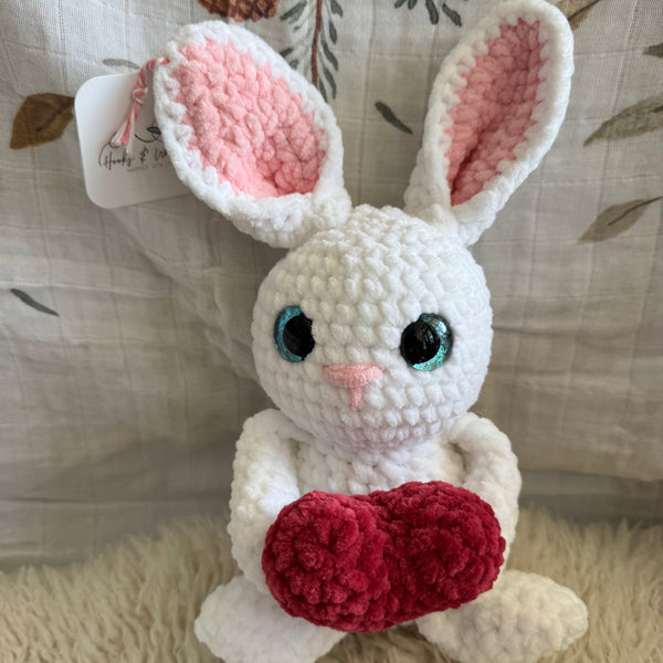 Crocheted bunny toy with pink ears and pink nose holding a heart on a textured fabric background