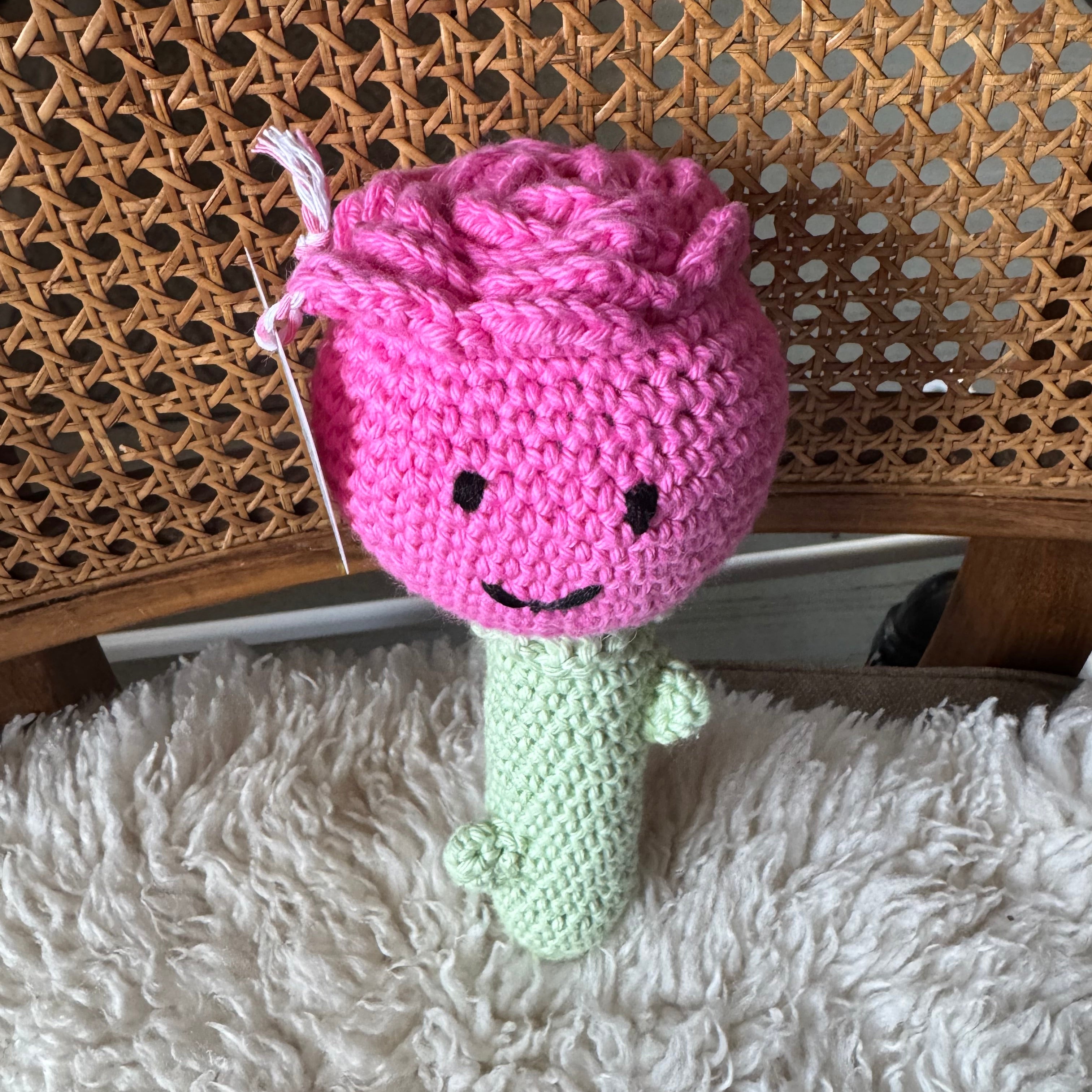 Rose crocheted toy with a face on a wooden surface next to a fluffy white blanket.