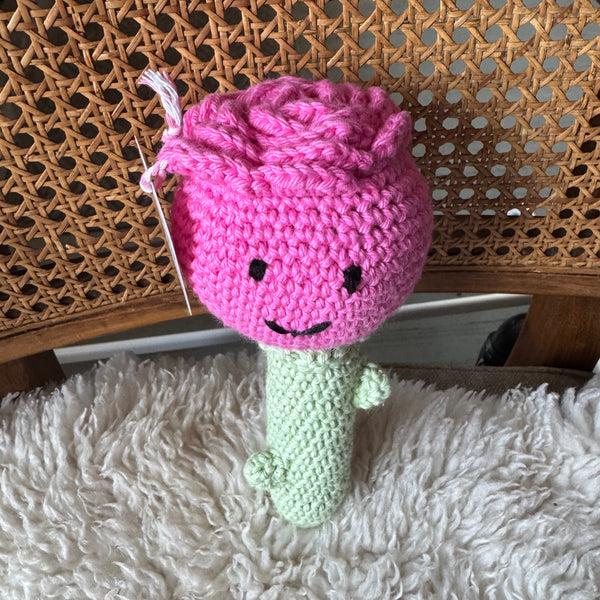 Rose crocheted toy with a face on a wooden surface next to a fluffy white blanket.