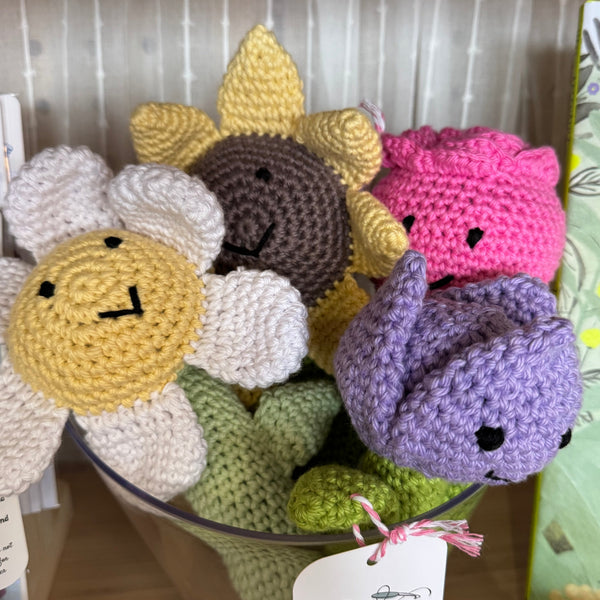 Collection of colorful crocheted flowers in a basket with a tag on a wooden surface.